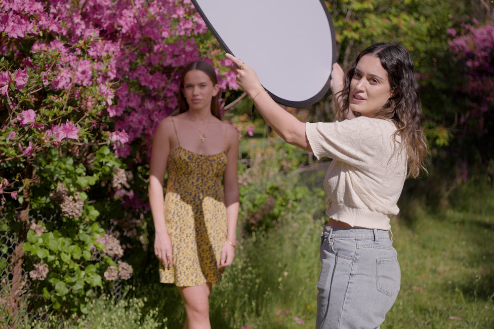 Julia Trotti holding a reflector in a photography scene. There is a model in a floral dress beside pink flowers in harsh midday sun